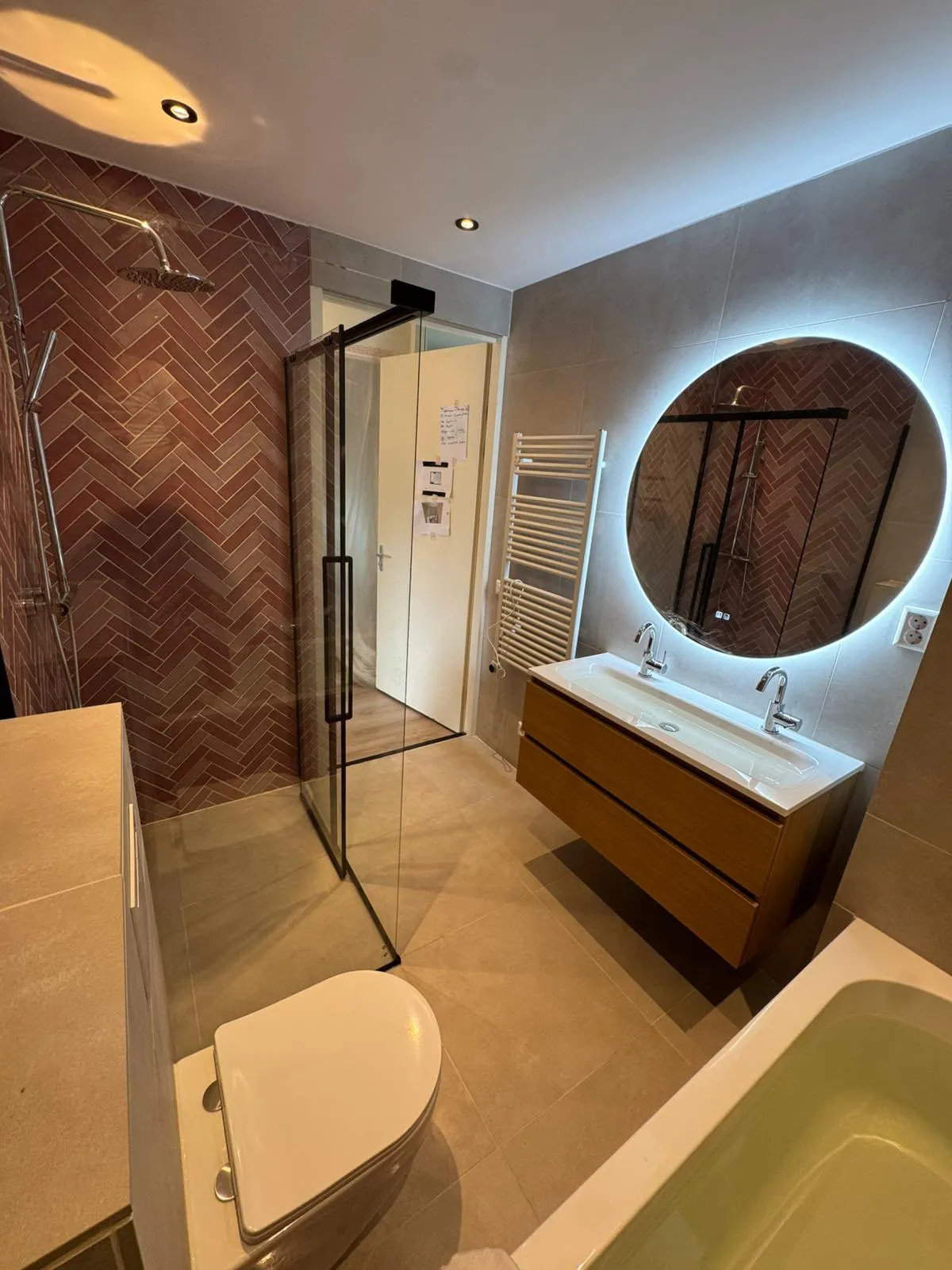 Badkamer met roze visgraattegels, glazen douchewand en ronde ledspiegel